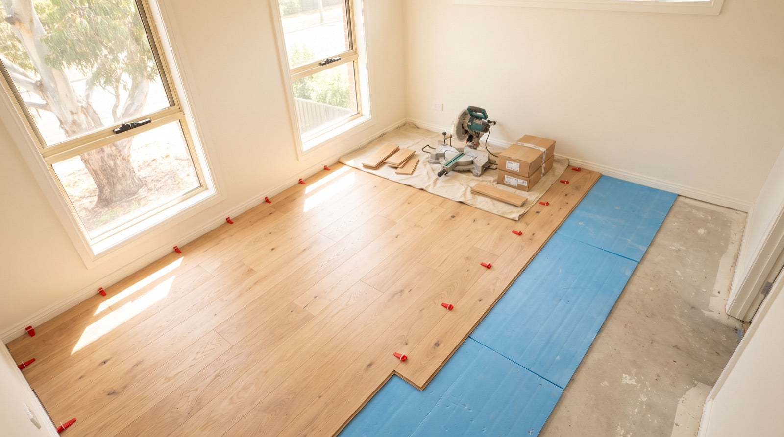 Click-lock floating floorboards part-laid across a bright Australian bedroom with spacers along the skirting line and a hire saw on a drop sheet